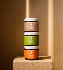 Stacked trio of Elija’s Nature body butter jars: Unscented, Lemon Blossom, and Mango Papaya on a pedestal against a beige-toned wall