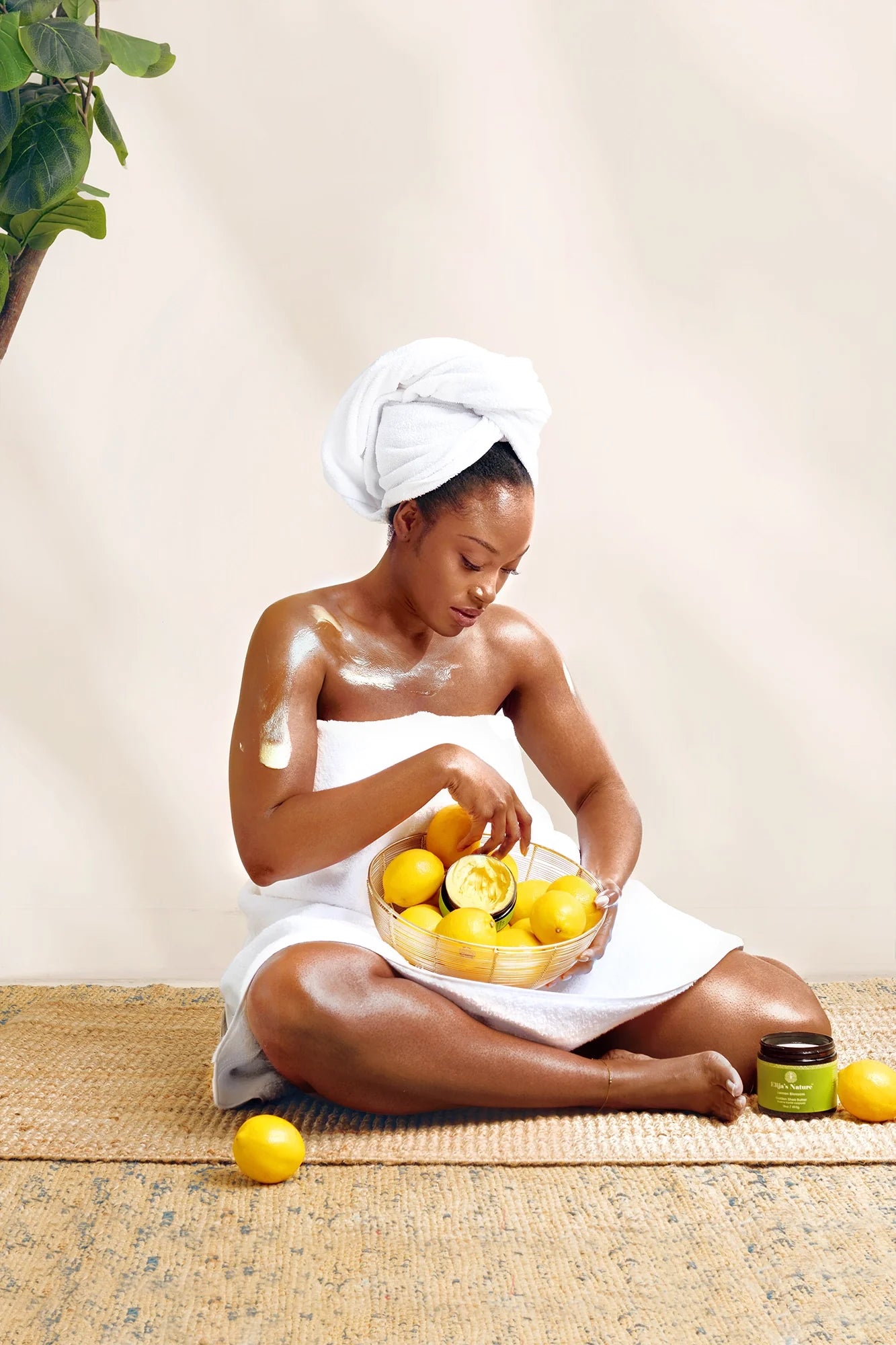 Golden Body Butter - #scent_Lemon Blossom #size_16oz -  A woman seated cross-legged in a towel applies body butter while holding a bowl of fresh lemons, conveying freshness and citrus-infused skincare