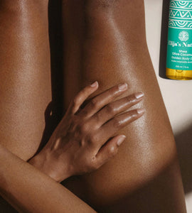 #scent_Olive Coconut #size_8oz - Woman with glowing skin sitting next to an Elija's Nature Golden Beauty Oil in the scent Olive Coconut